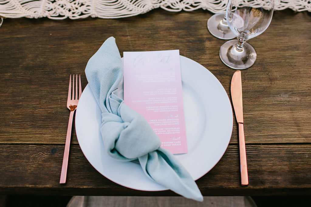 wedding reception table setting with knotted blue napkin and rose gold silverware with neon ombre pink menu card at Triunfo Creek Vineyards