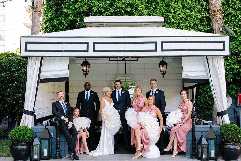 A glam and California infused wedding at Viceroy Santa Monica, wedding party photo with bridesmaids in pink slip dresses and feathered fans, groomsmen in navy suits