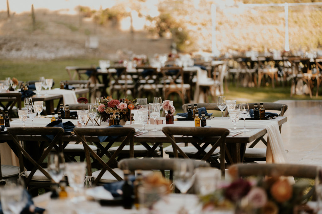 A whimsical wedding reception at Triunfo Creek Vineyards, rustic wood cross back chairs with navy blue napkins and pink and orange floral centerpieces