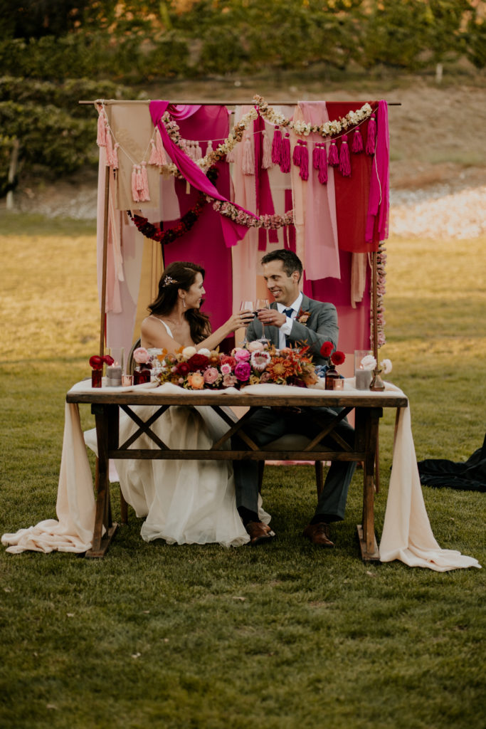 A whimsical wedding reception at Triunfo Creek Vineyards, bride and groom sweetheart table with pink fabric and tassel arch