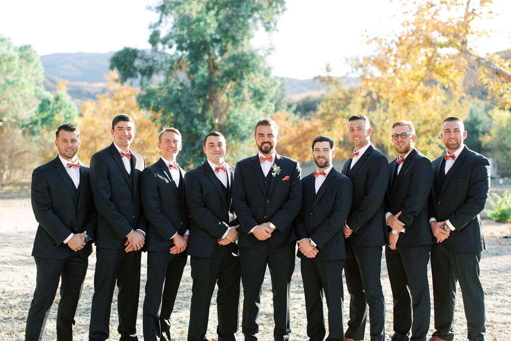 Groom with groomsmen in black suit with rust colored tie getting ready for wedding day