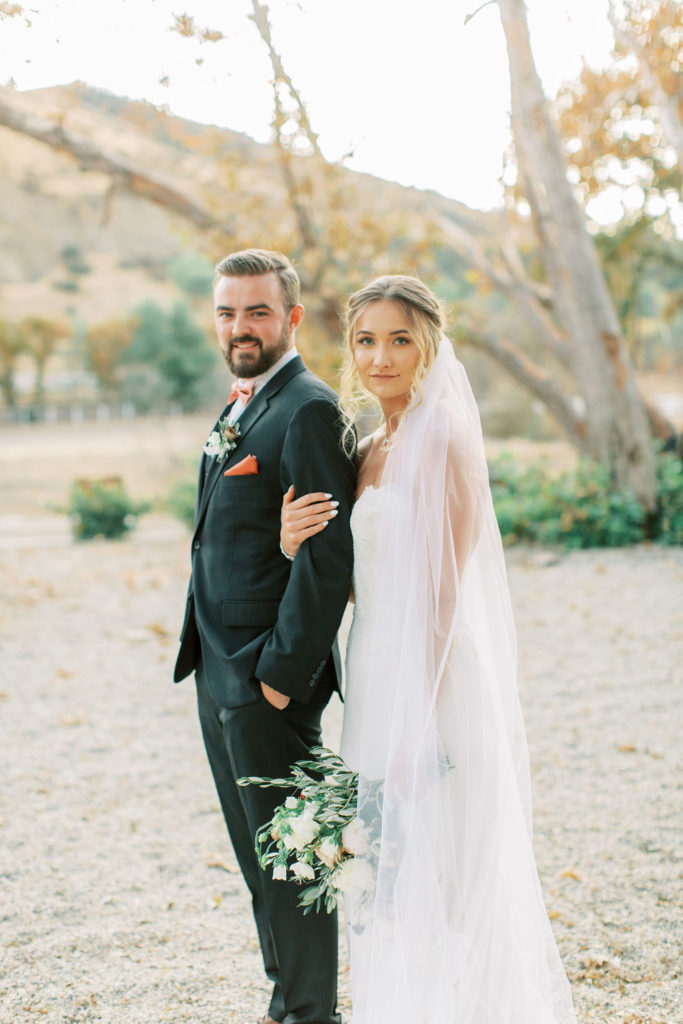 bride in lace wedding dress and cathedral length veil with groom in black suit and rust colored tie take a portrait shot in vineyard