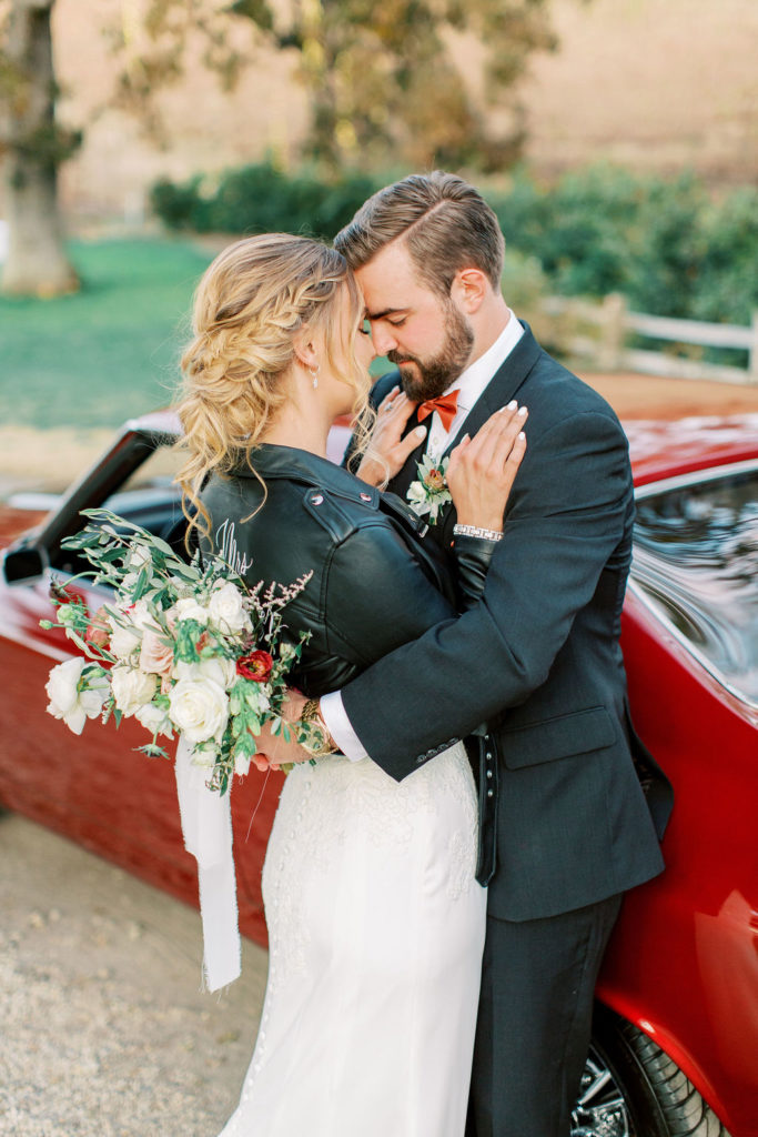 bride in lace wedding dress and custom last name leather jacket with groom in front of red vintage mustang car