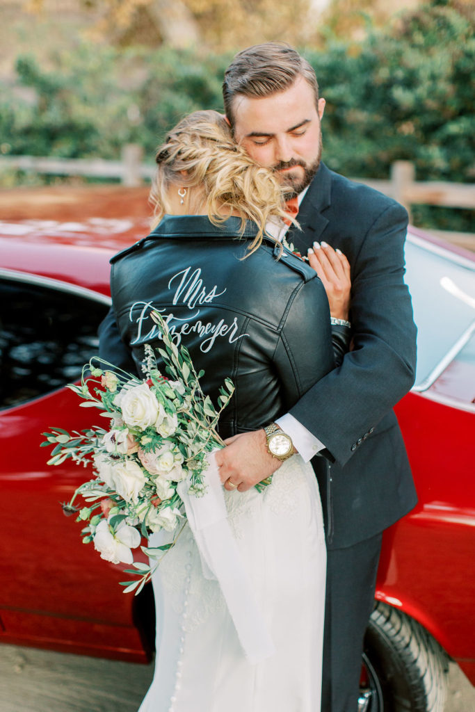 bride in lace wedding dress and custom last name leather jacket with groom in front of red vintage mustang car