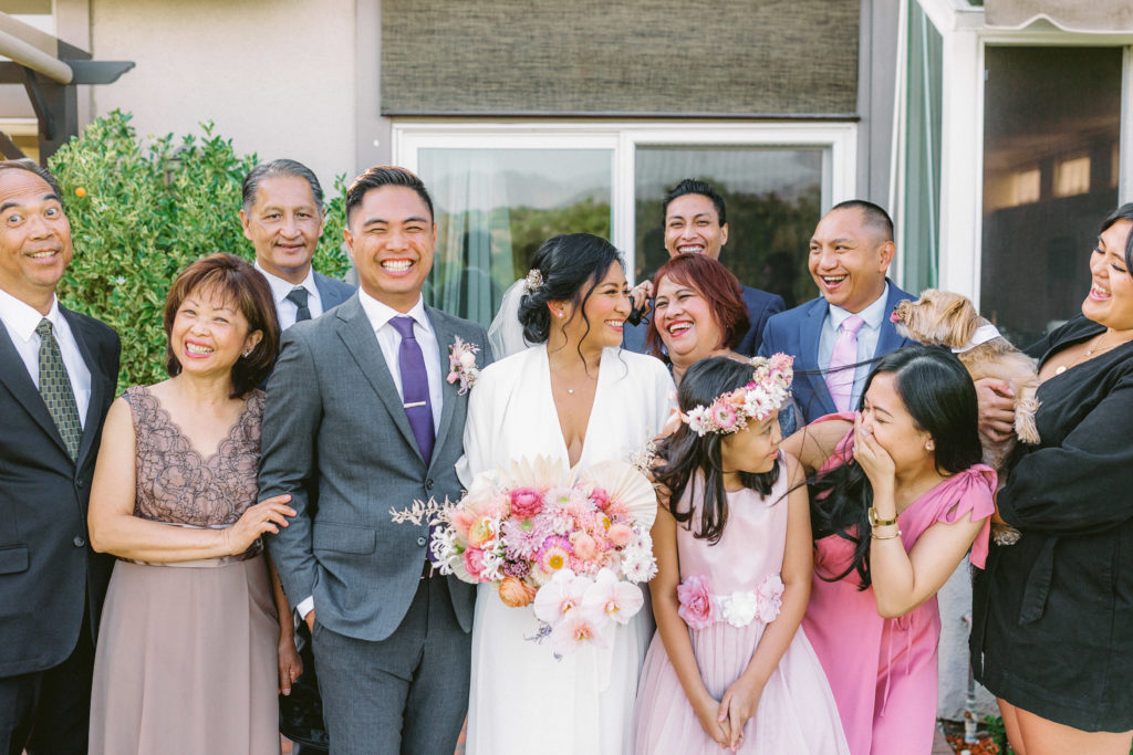 bride wearing Reformation wedding dress and groom with grey suit and purple tie portrait shot with family during small backyard ceremony