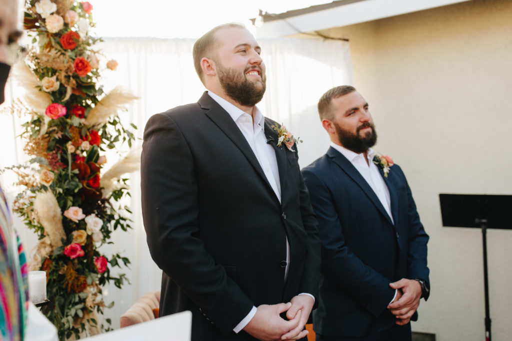 A groom watches his bride walk down the aisle during an intimate backyard wedding