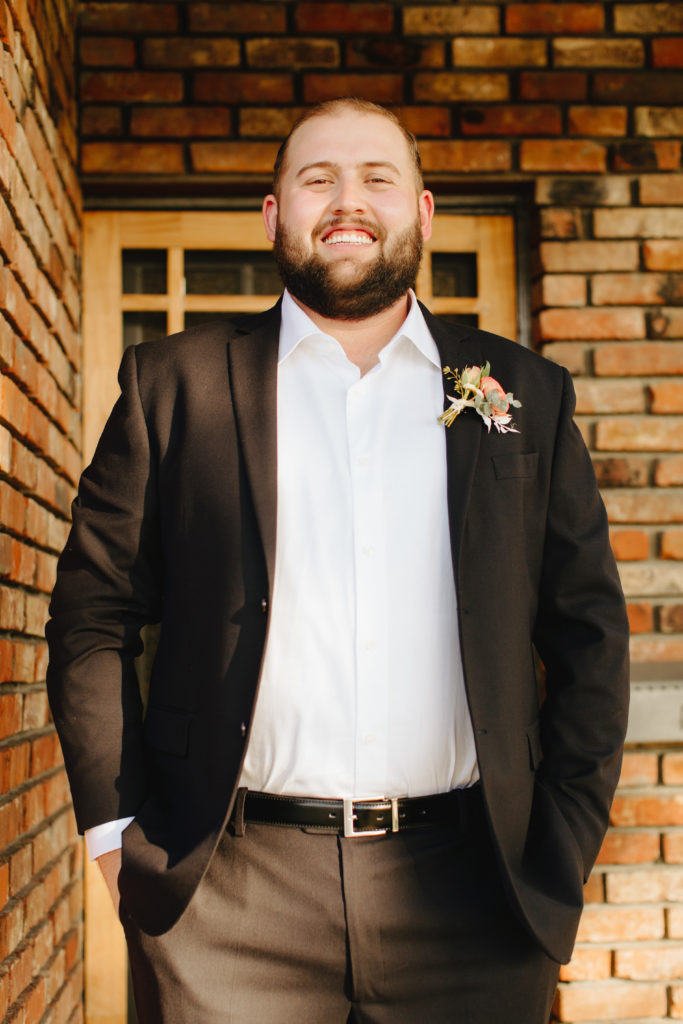 Groom wearing classic black blazer with no tie poses on front porch before intimate backyard wedding