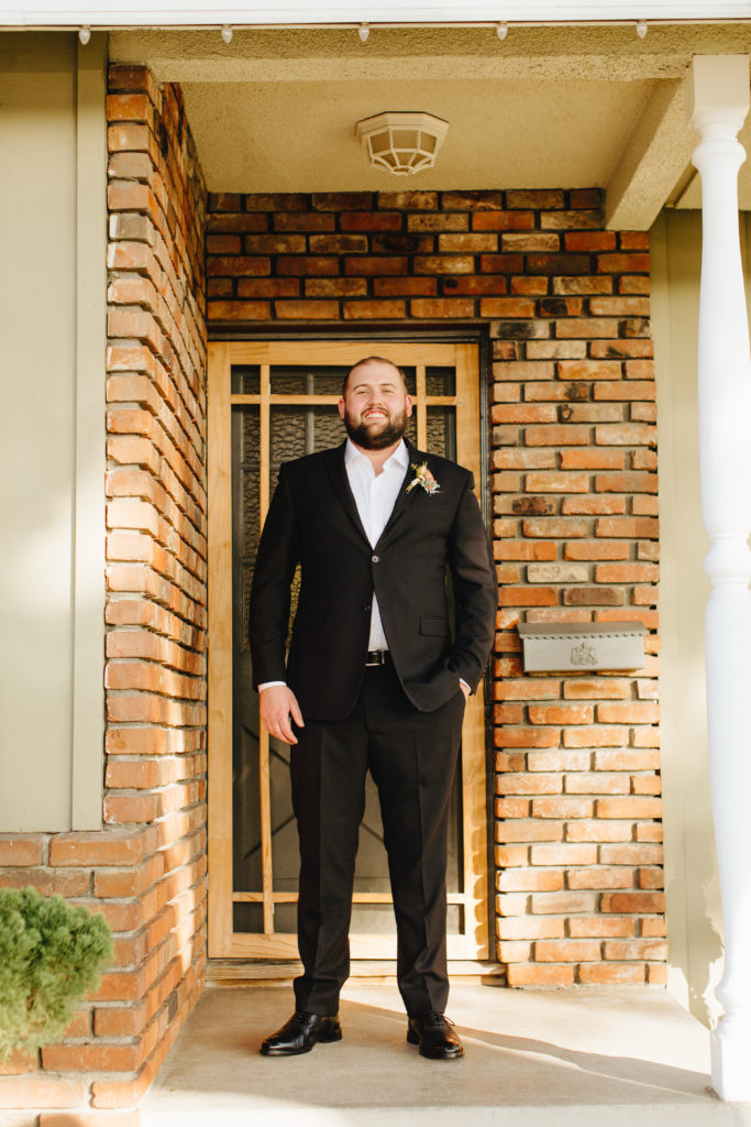 Groom wearing classic black blazer with no tie poses on front porch before intimate backyard wedding