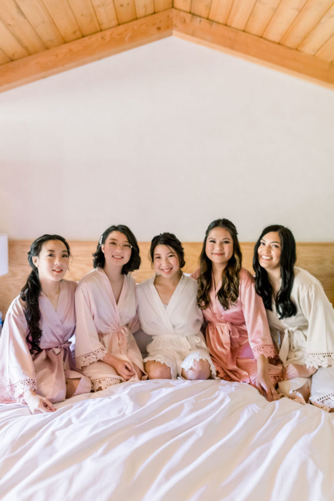 bride in satin robe sits on bed with bridesmaids in pink coral and beige satin robes