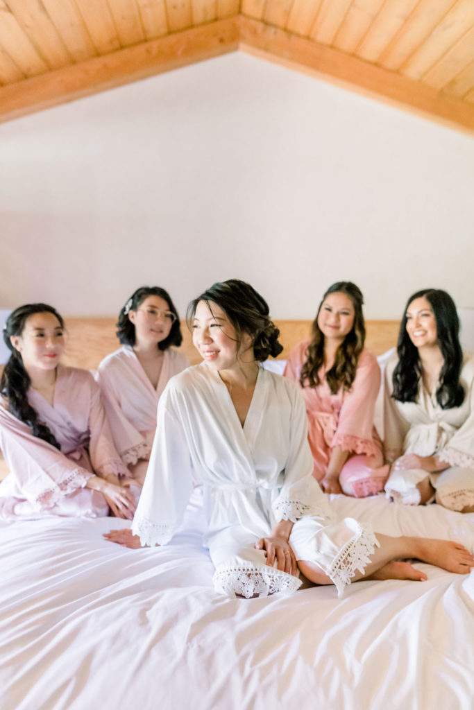 bride in satin robe sits on bed with bridesmaids in pink coral and beige satin robes