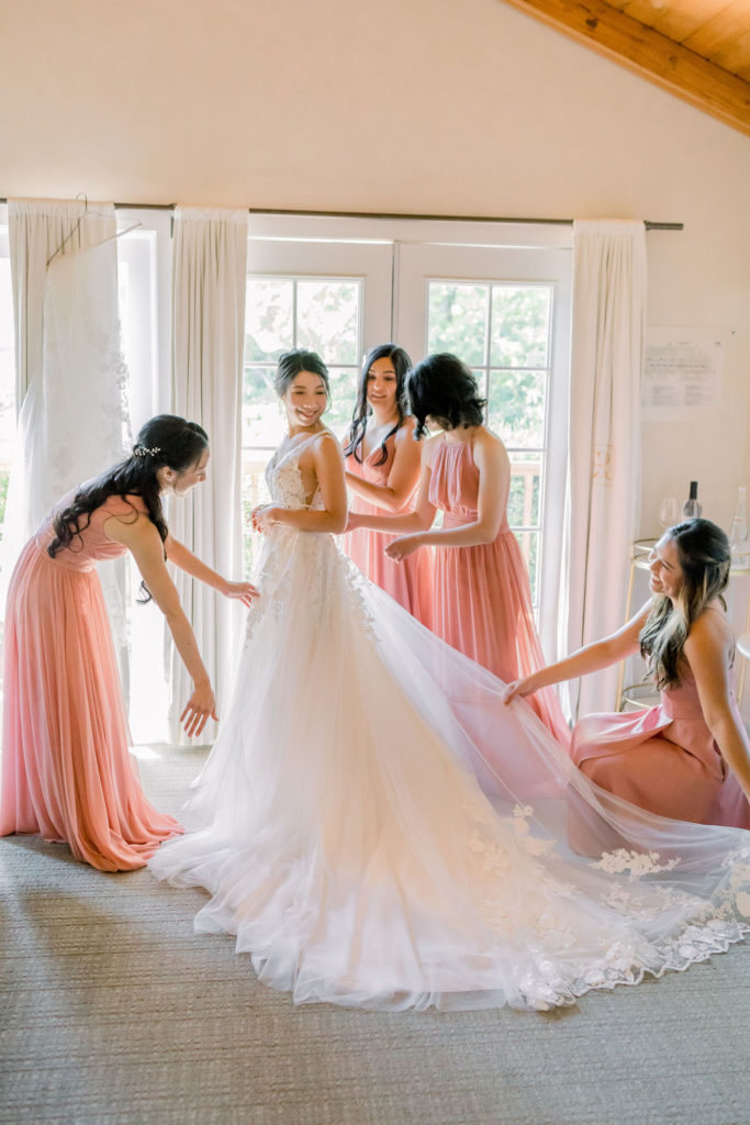 bridesmaids in coral pink bridesmaid dresses helps bride into her wedding dress