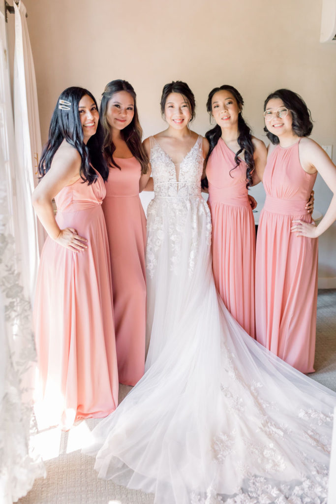 bride in a deep v-neck wedding dress with floral appliqué stands with bridesmaids wearing pink coral bridesmaid dresses
