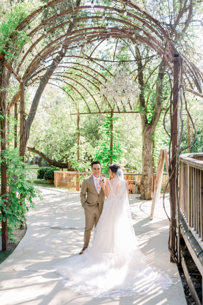 An enchanting wedding at Calamigos Ranch, groom in tan suit with floral tie has walks with bride wearing a floral lace appliqué deep v-neck wedding dress and cathedral length veil