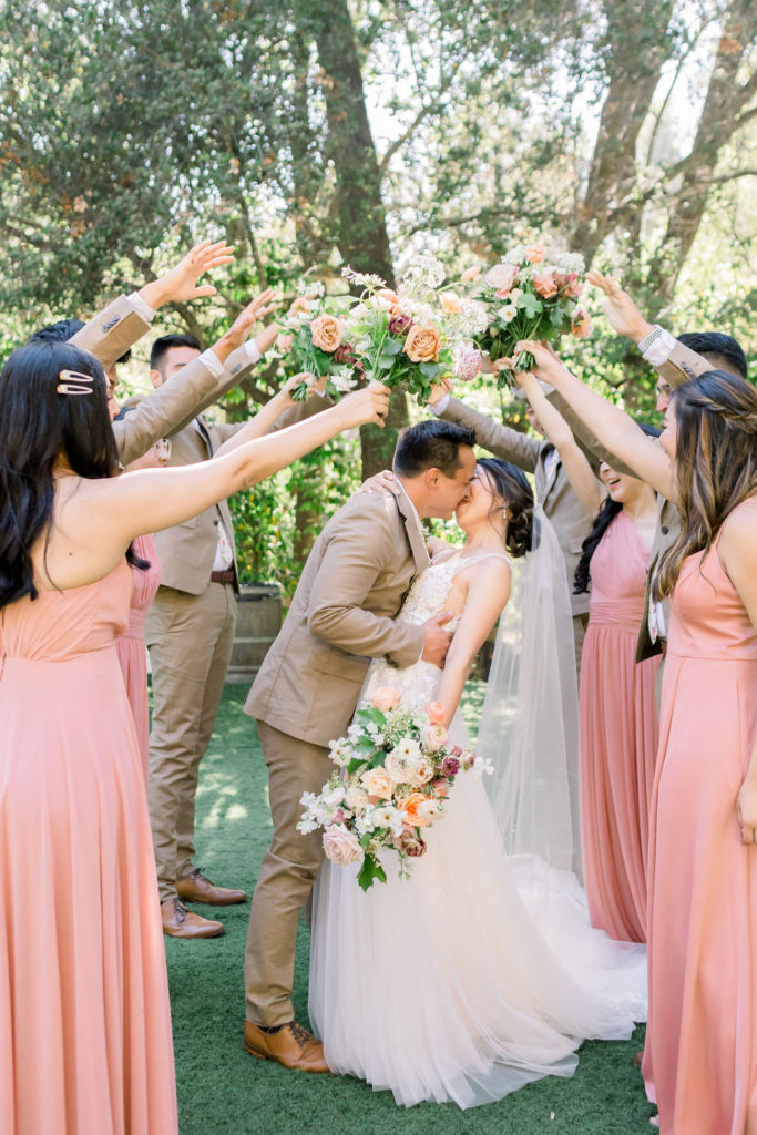 groom and bride kiss under flower bridge with wedding party wearing tan suits with floral ties and coral pink bridesmaid dresses