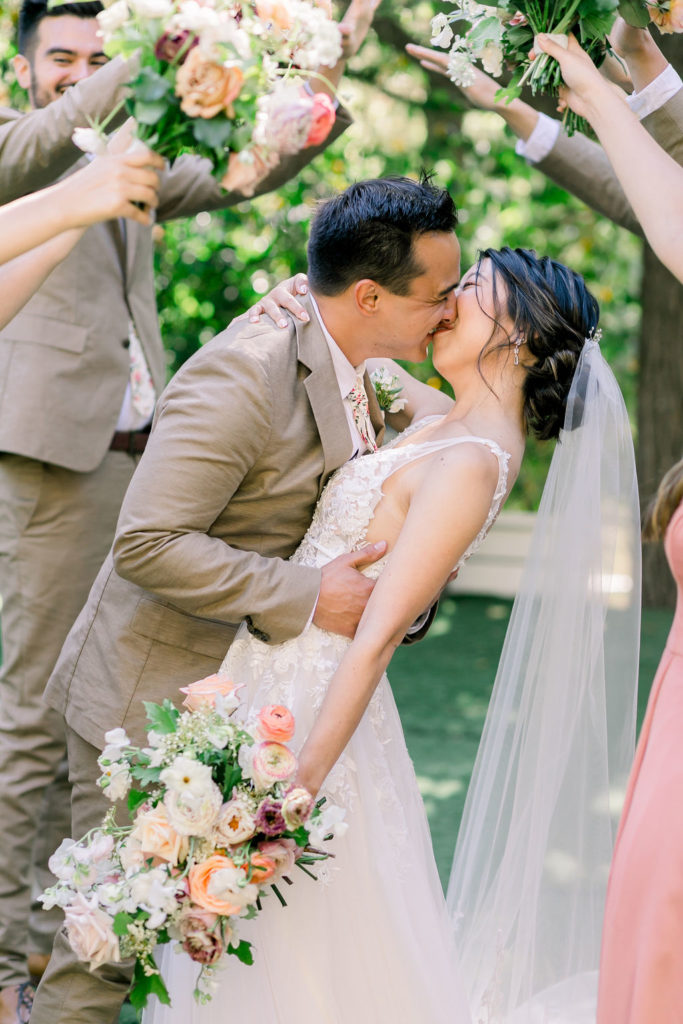 groom and bride kiss under floral bridge with wedding party wearing tan suits with floral ties and coral pink bridesmaid dresses