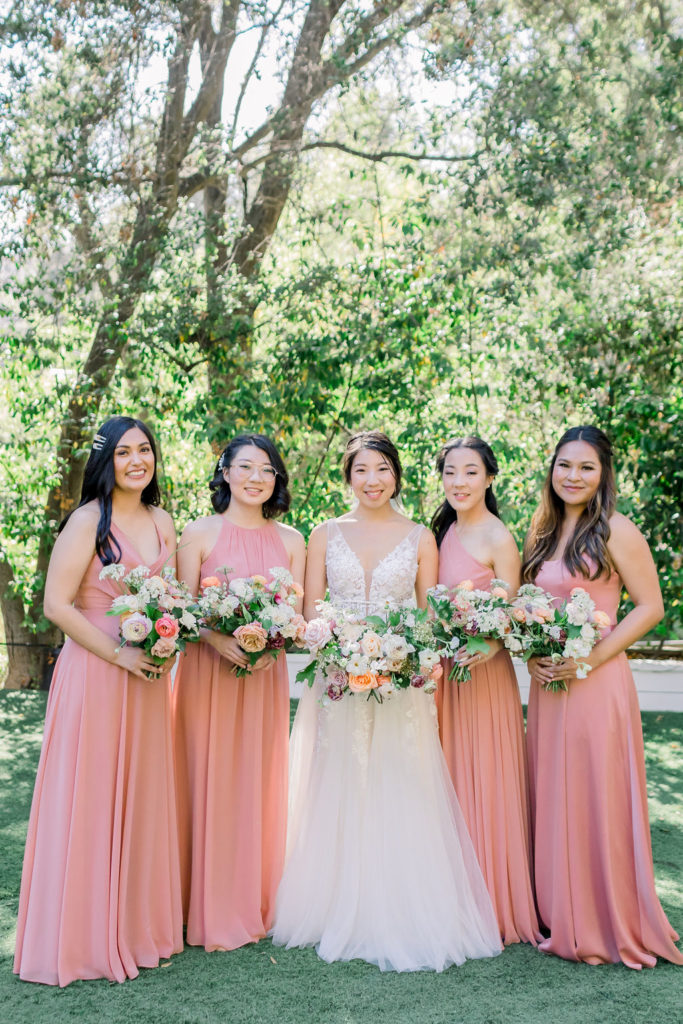 bride wearing deep v wedding dress with floral appliqué stands with bridesmaids wearing coral pink bridesmaid dresses