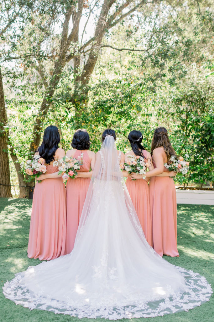 bride wearing deep v wedding dress with floral appliqué stands with bridesmaids wearing coral pink bridesmaid dresses