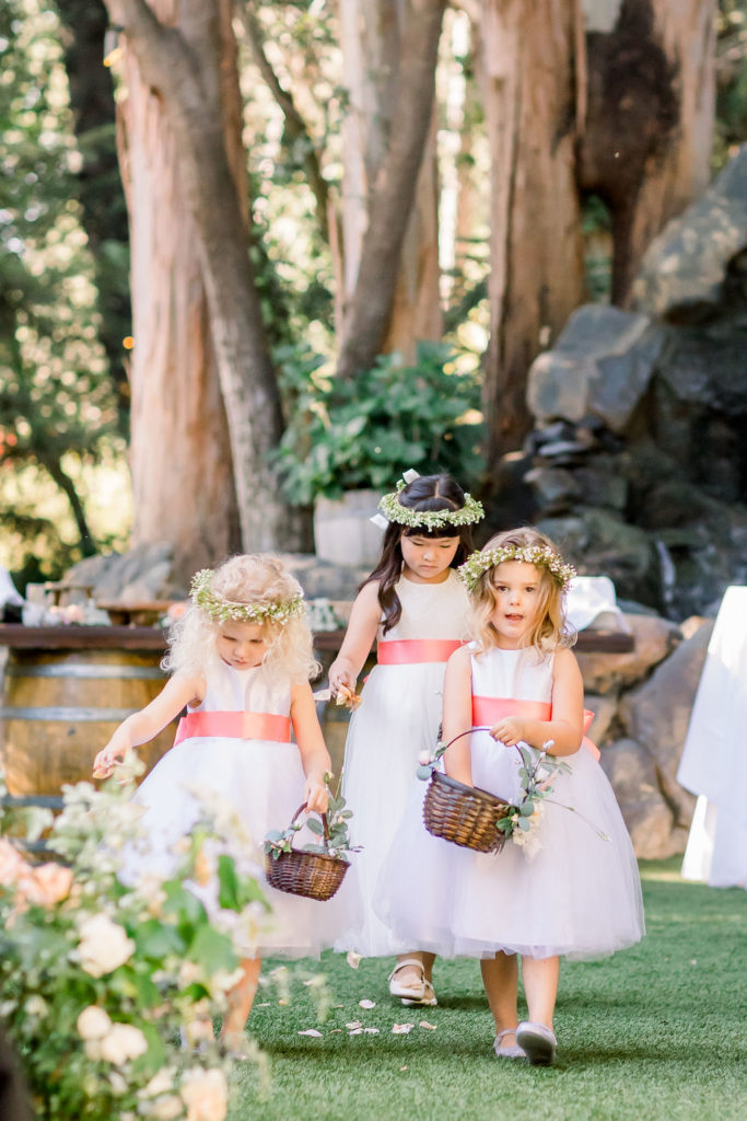 flower girls wearing matching white dresses and pink sashes and flower crowns place petals down wedding aisle