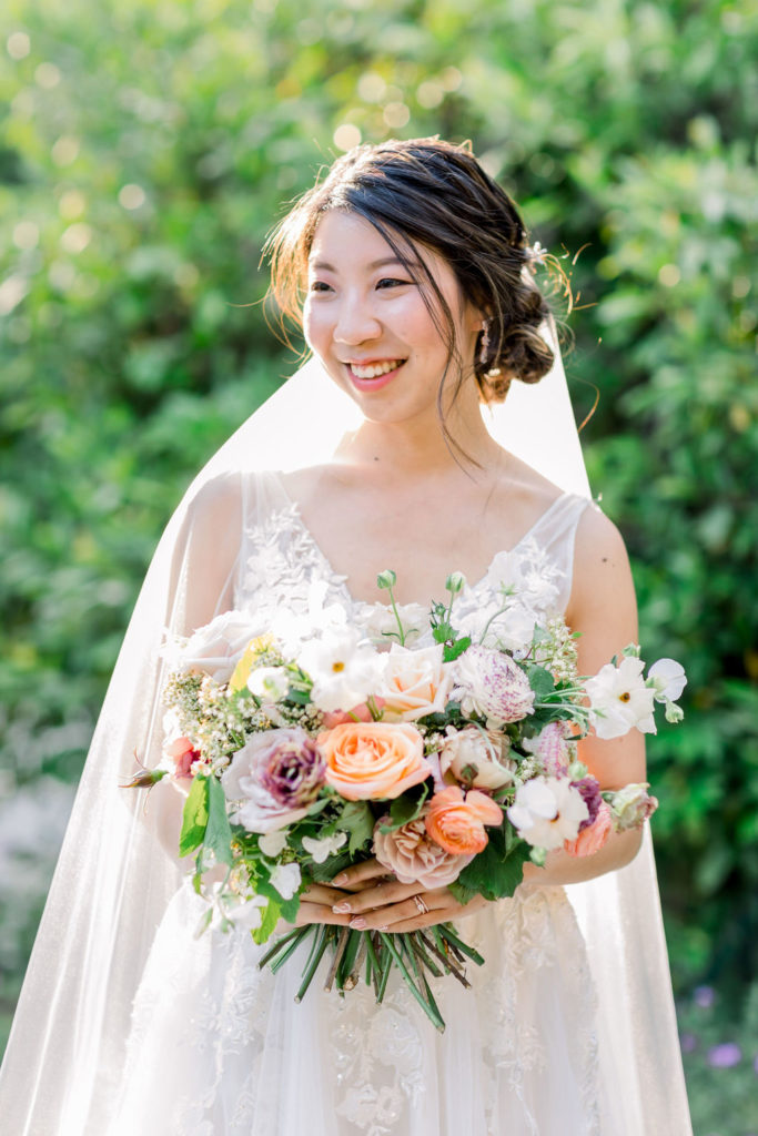 bride wearing a floral lace appliqué deep v-neck wedding dress and cathedral length veil while holding a bridal bouquet of orange, pink, beige, grey and green