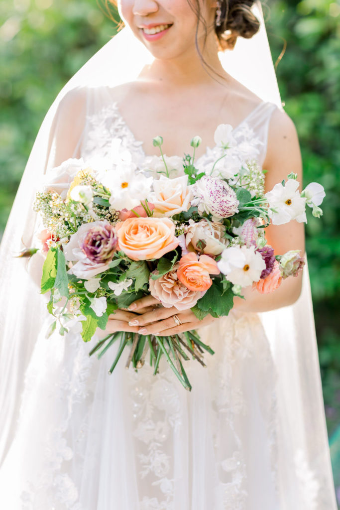 bride wearing a floral lace appliqué deep v-neck wedding dress and cathedral length veil while holding a bridal bouquet of orange, pink, beige, grey and green