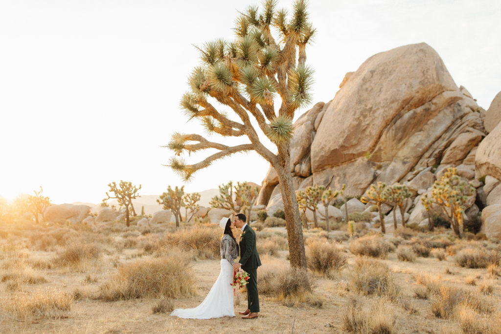 bohemian bride wearing distressed denim jack and hat with groom in a dark suit and pink bowtie take portraits during sunset after desert elopement in Joshua Tree National Park