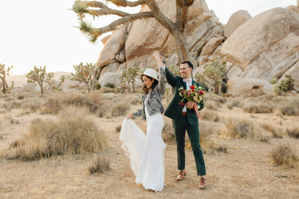 bohemian bride wearing distressed denim jack and hat with groom in a dark suit and pink bowtie take portraits during sunset after desert elopement in Joshua Tree National Park
