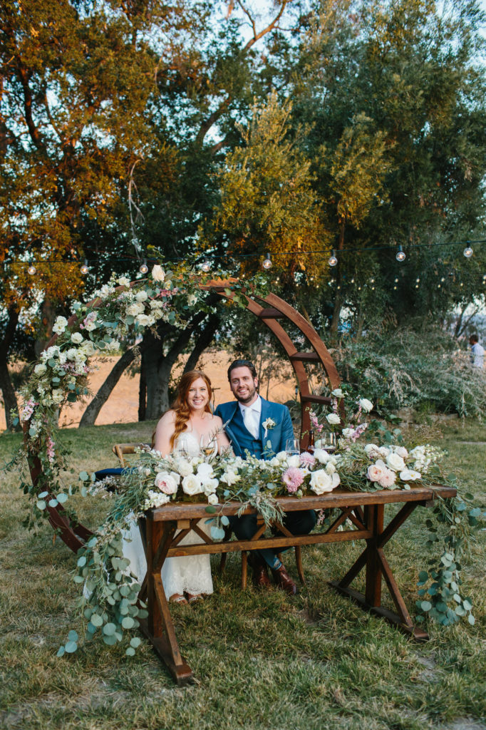 bride and groom sit at sweet heart table with circle arch from ceremony behind them