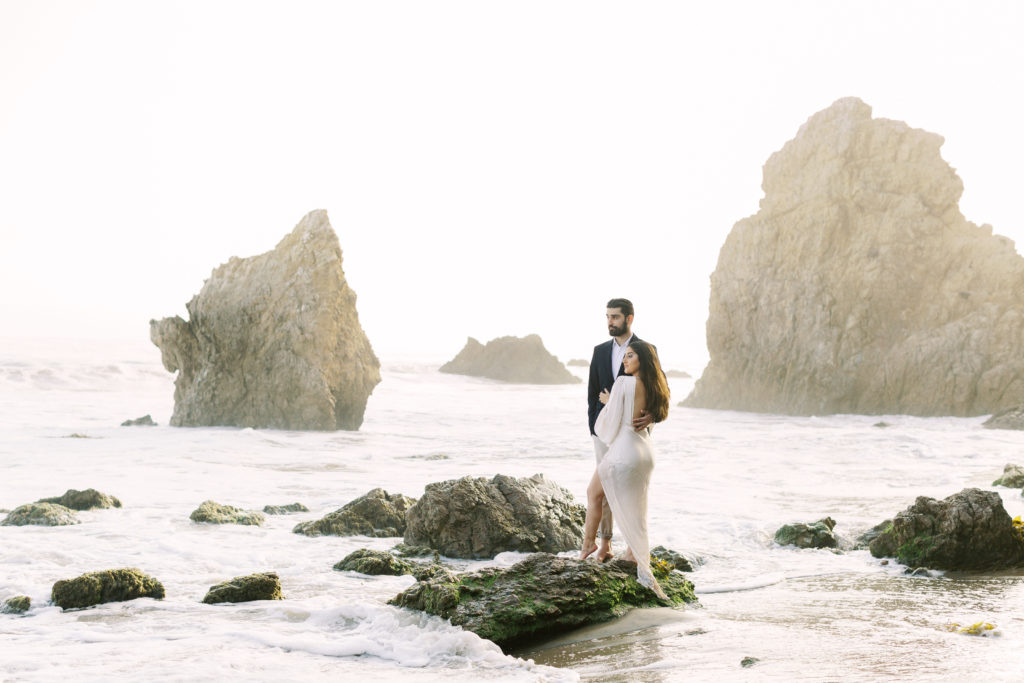 bride to be in white deep v neck dress with groom to be in black suit on beach engagement shoot