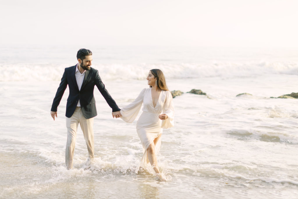bride to be in deep v neck white dress walks with groom to be in black suit jacket and khaki pants in the waves for their beach engagement shoot