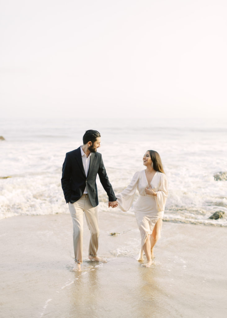 bride to be in white deep v neck dress with groom to be in black suit on beach engagement shoot