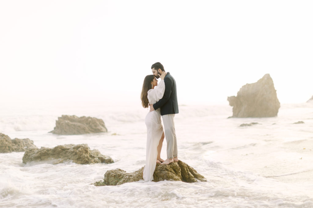 couple stand on rocks in the ocean during their engagement photos on the beach