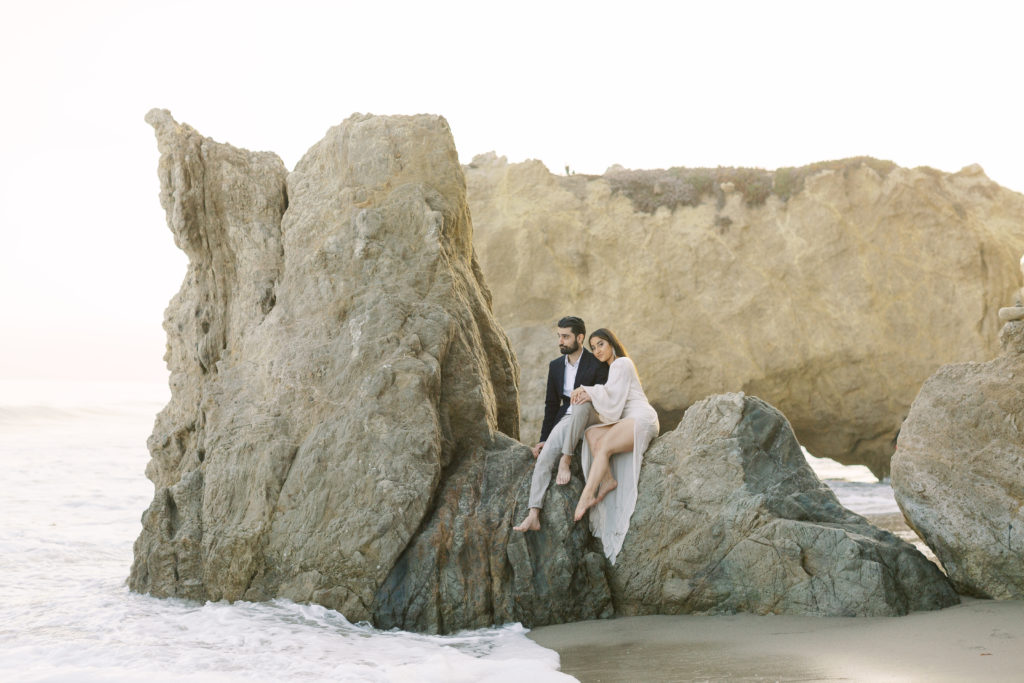 couple sits on rock during beach engagement photo shoot