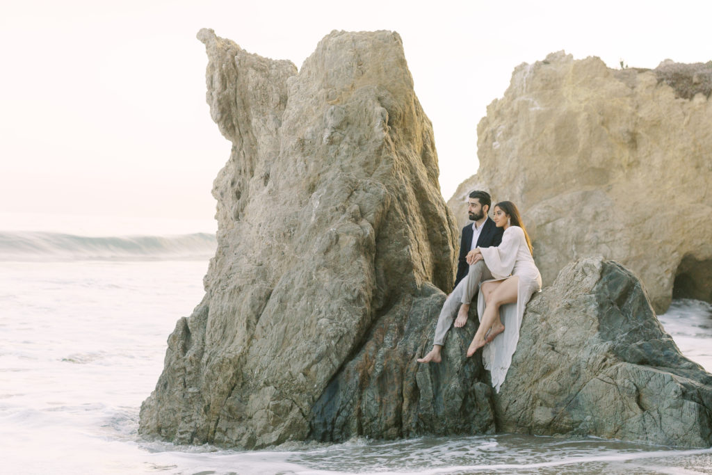 bride to be in white deep v neck dress with groom to be in black suit sit on rocks on a beach