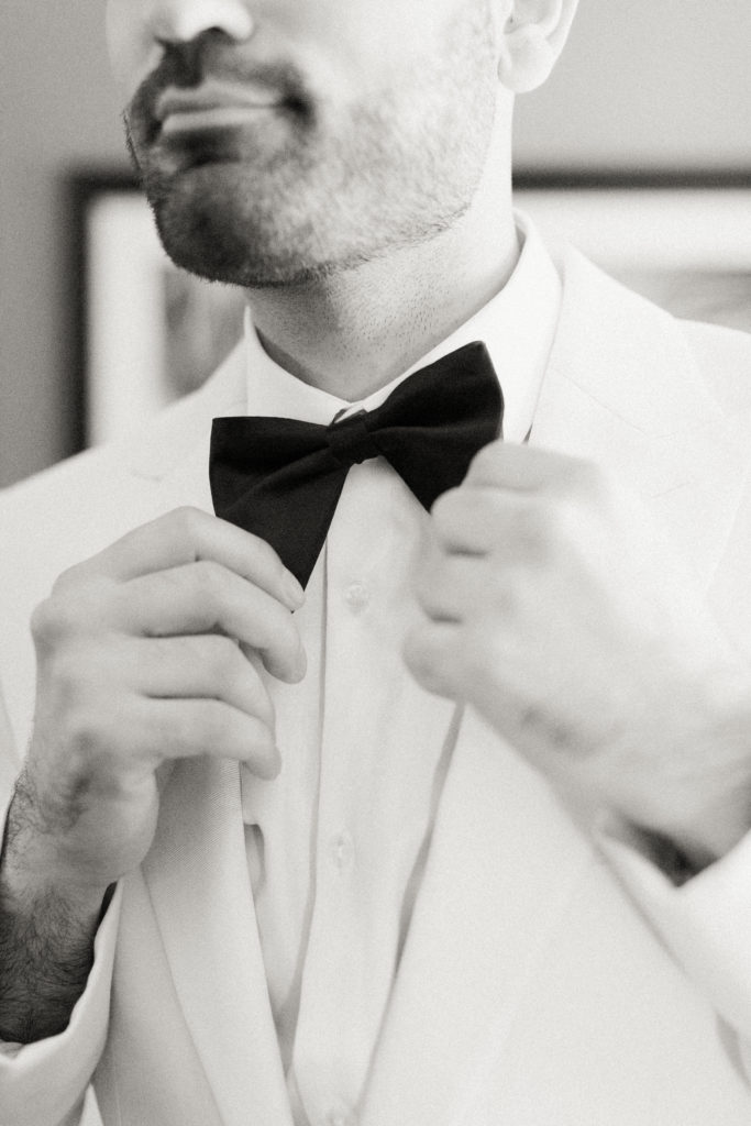 groom in white tuxedo and black bowtie gets ready for wedding