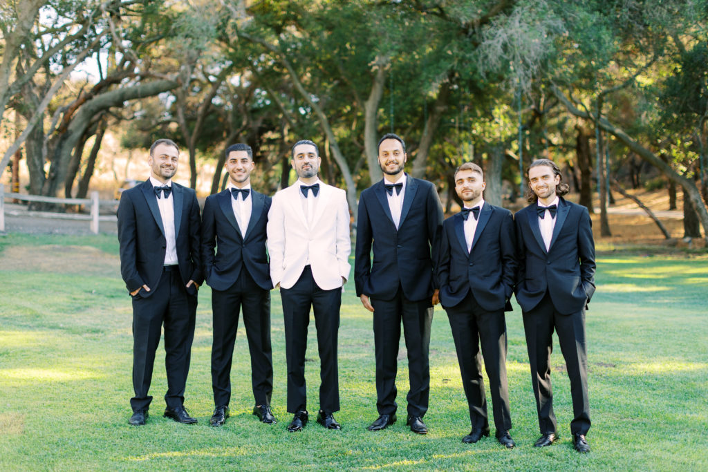 groom in white suit jacket stands with groomsmen in black suits with bowties