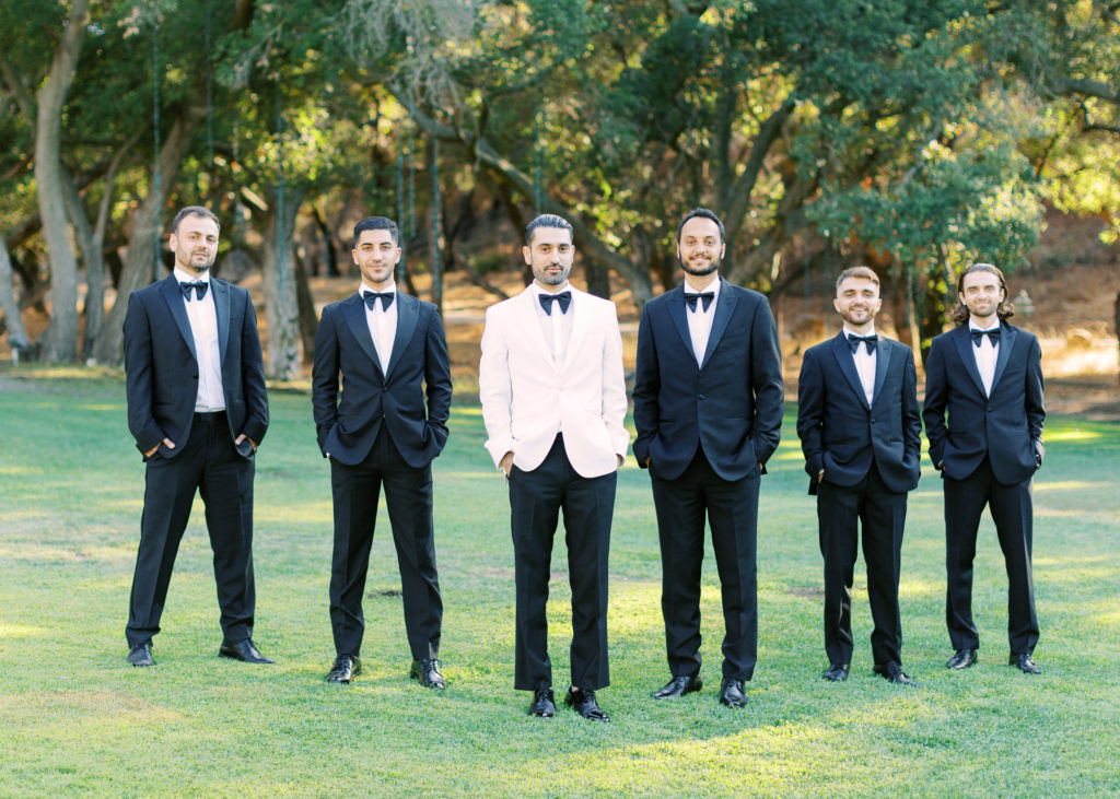 groom in white suit jacket stands with groomsmen in black suits with bowties