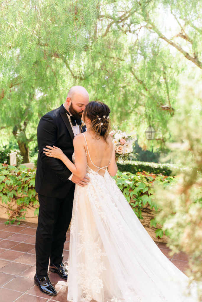 bride with open back dress kisses groom wearing black tuxedo suit