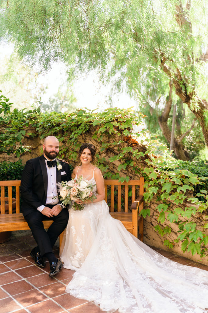groom in classic black tux sits with bride in floral appliqué dress on bench during portraits