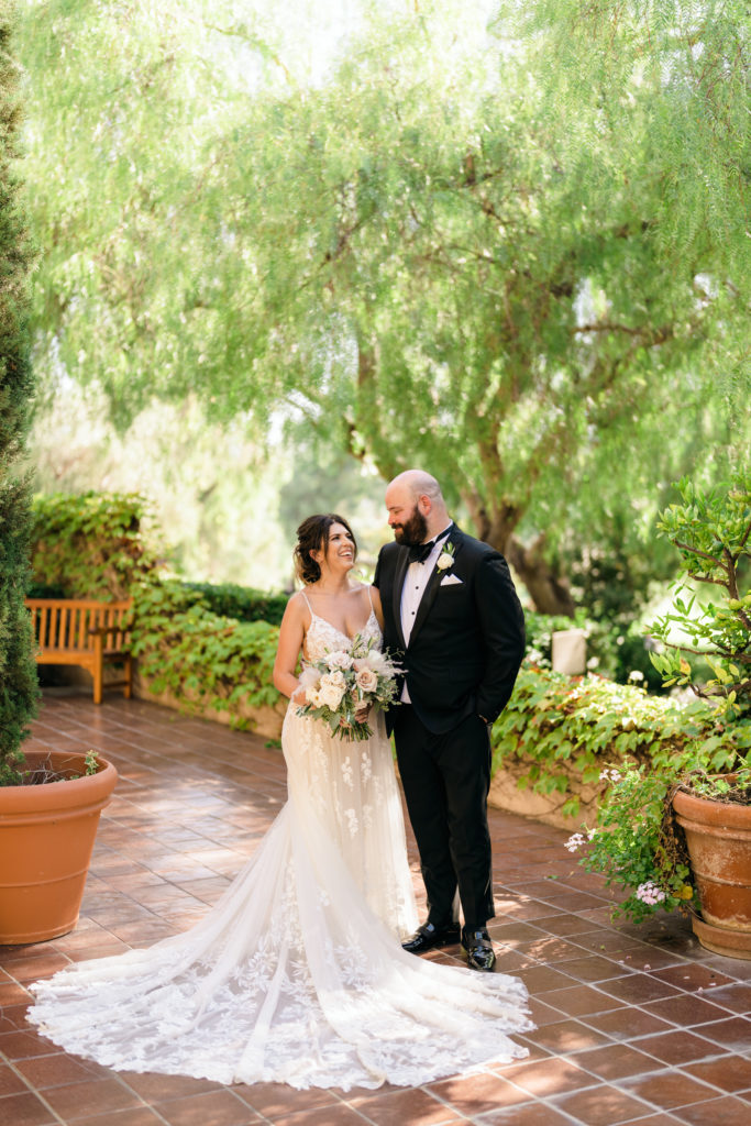 bride in floral appliqué dress with long train stands with groom in black tuxedo suit on garden terrace of Ranch Bernardo Inn