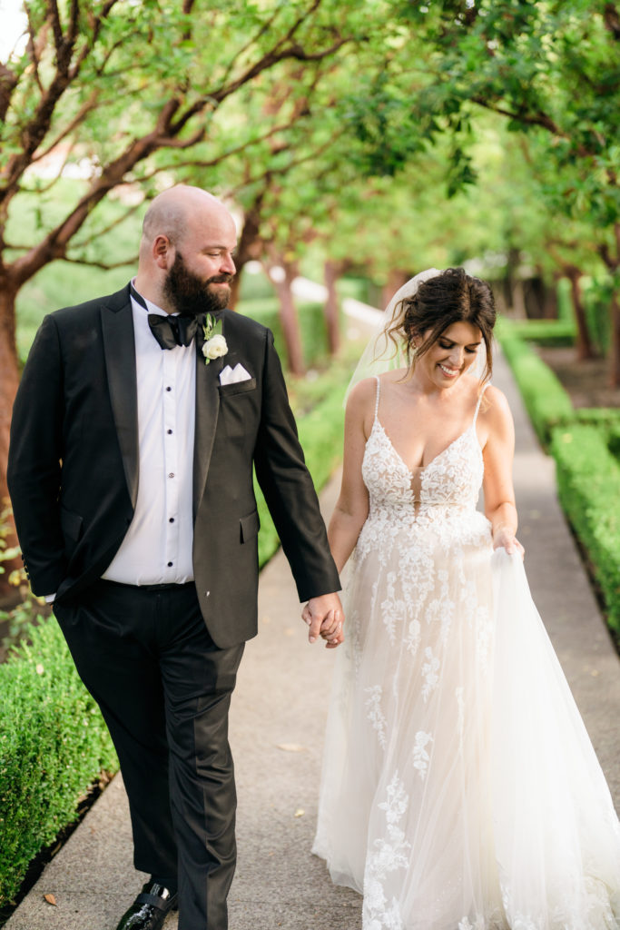groom in black suit tuxedo holds hands with bride in floral appliqué dress in garden