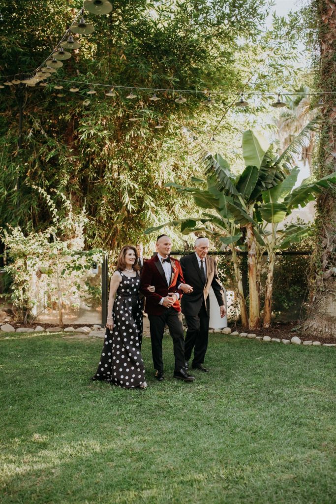 groom in red velvet suit jacket walks with parents down the aisle