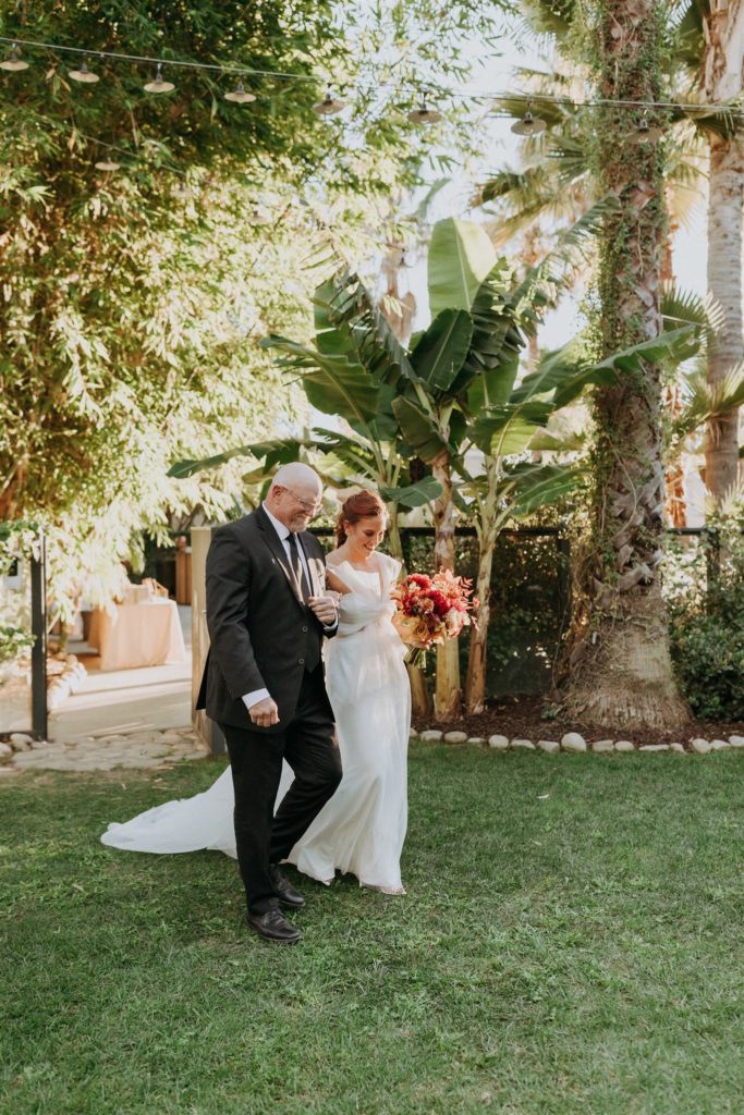 bride wearing modern asymmetrical wedding dress and bright bridal bouquet walks down the aisle with her father