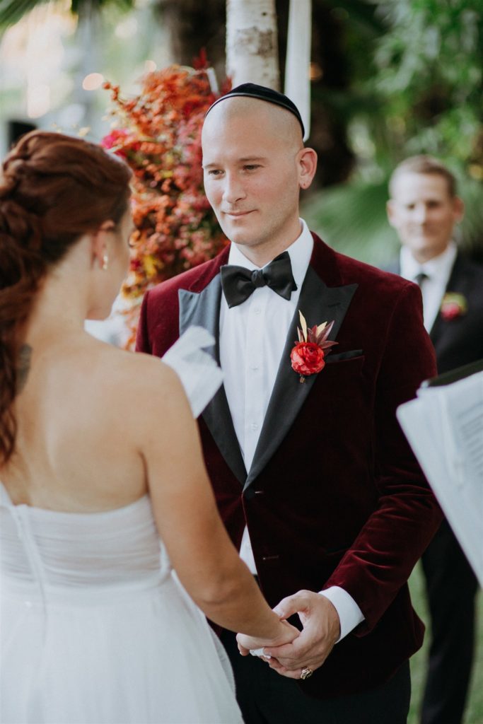bride in modern asymmetrical dress stands with groom in red velvet suit jacket during mid-century wedding ceremony at Capri Hotel in Ojai