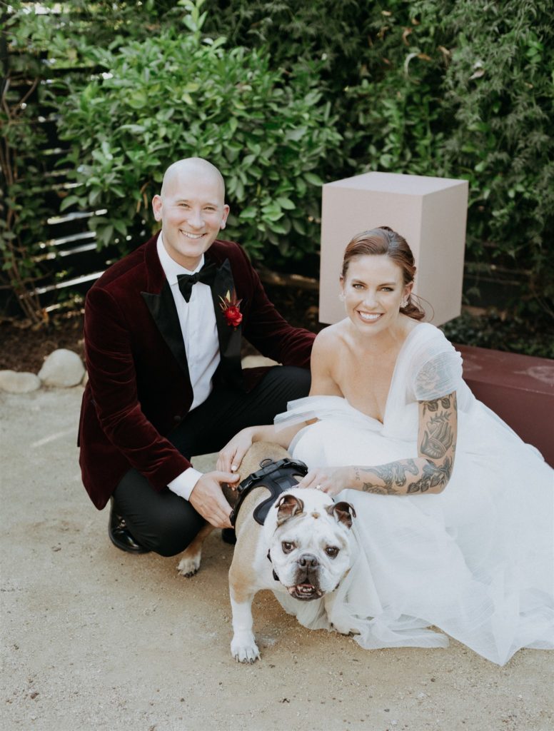 bride in modern asymmetrical dress stands with groom in maroon velvet suit jacket and their dog in the Capri Hotel in Ojai
