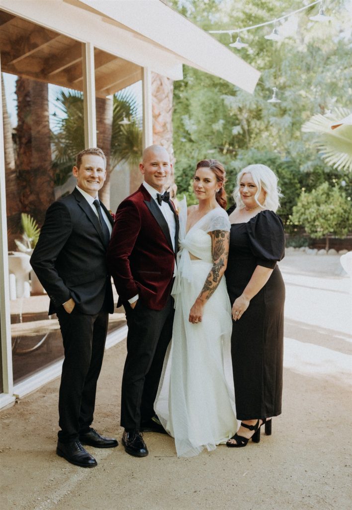 bride in modern asymmetrical dress and groom in maroon velvet suit jacket stand with their wedding party in a black suit and dress
