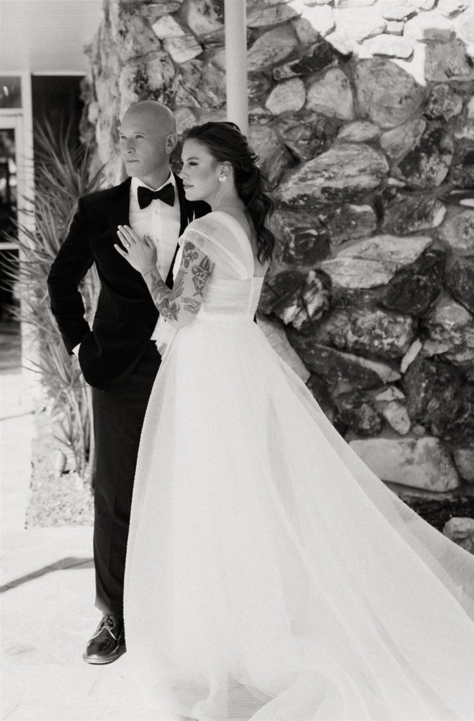 bride in modern asymmetrical dress stands with groom in maroon velvet suit jack in front of the Capri Hotel in Ojai