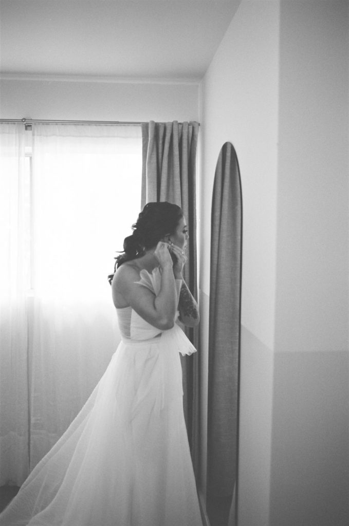 bride in asymmetrical modern wedding dress puts earrings on in mirrorr