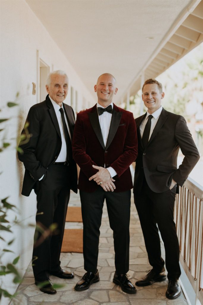 groom in maroon velvet suit stands with groomsman and father