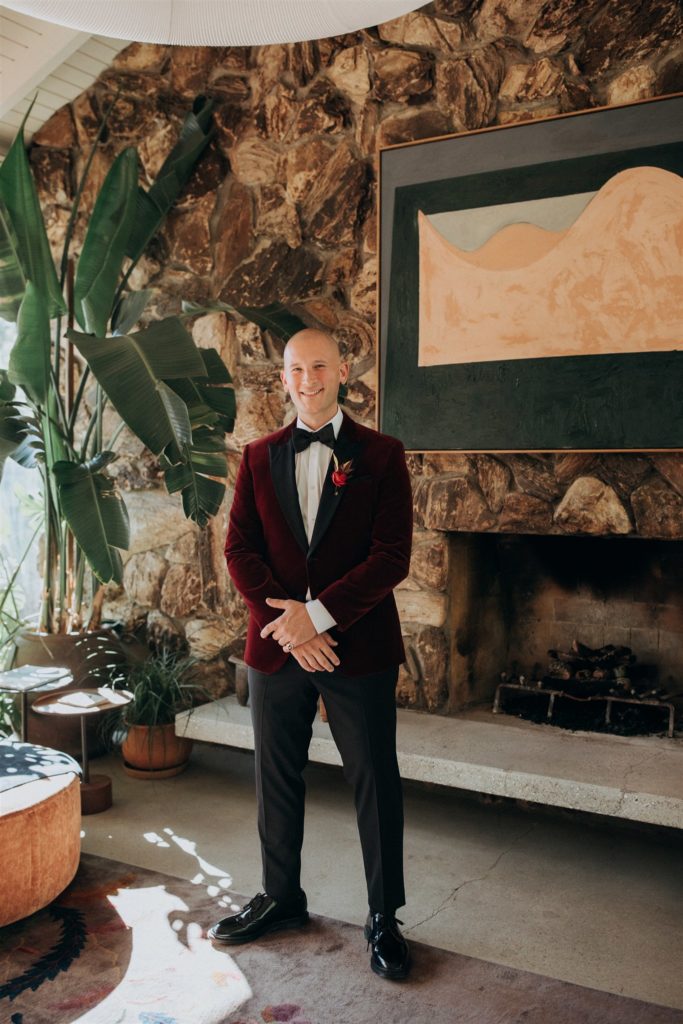 groom in maroon velvet suit jacket and black bowtie stands in mid-century lobby of Capri Hotel