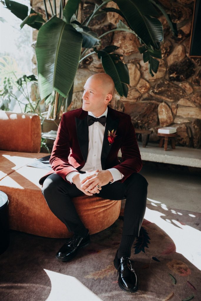 groom in maroon velvet suit jacket and black bowtie poses in mid-century lobby of Capri Hotel